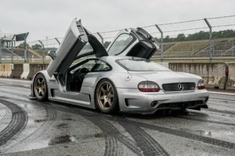 Mercedes-Benz CLK GTR argentée vue en grand angle au ras du sol sur une ligne de stands mouillée, portière en ciseaux ouverte, atmosphère circuit historique années 90